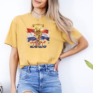 May include: Mustard yellow t-shirt featuring a graphic of the Croatian flag, a gold trophy, and the text "2026". The word "Croatia" is printed above the trophy. The shirt has short sleeves and a crew neck.