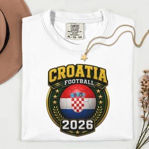 Peut inclure: T-shirt blanc avec un motif de football croate. Le design pr&eacute;sente un ballon de football avec le drapeau croate, le mot "CROATIA" en or et l'ann&eacute;e "2026". Un collier en or avec un pendentif en forme d'&eacute;toile est pos&eacute; sur le t-shirt.