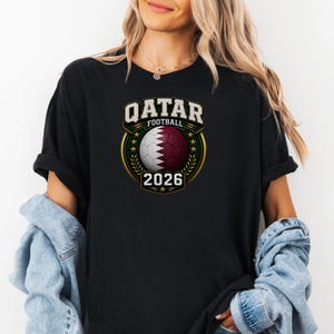 May include: Black t-shirt featuring a graphic of a soccer ball with the Qatar flag design, the text "QATAR FOOTBALL" above, and "2026" below. The design is framed by a gold laurel wreath. The shirt has rolled-up sleeves.