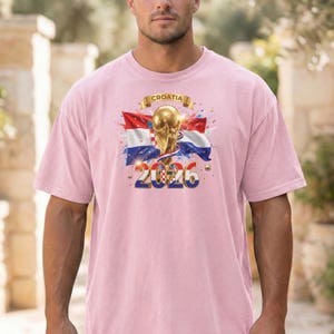 May include: A light pink t-shirt featuring a graphic design with the word "CROATIA" above a golden trophy, the Croatian flag, and the number "2026". The design is centered on the shirt.