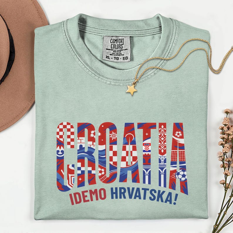 May include: A light green t-shirt with the word "CROATIA" in red, white, and blue patterned letters. The text "IDEMO HRVATSKA!" is below. A gold necklace with a star pendant and a brown hat are also visible.