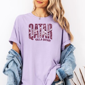 Puede incluir: Una camiseta de color lila claro con la palabra "QATAR" en una fuente decorativa granate. Debajo de la palabra "QATAR" est&aacute; la frase "YALLA QATAR!" en granate. La camiseta es de manga corta y cuello redondo.