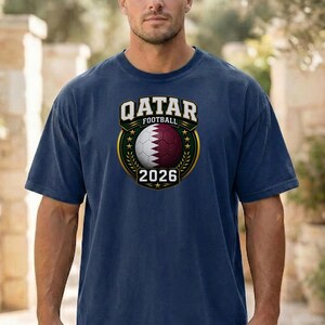May include: Navy blue t-shirt featuring a Qatar Football emblem. The emblem includes a soccer ball with the Qatar flag design, the text "QATAR FOOTBALL" and the year "2026". The emblem is surrounded by a gold border and green leaves.