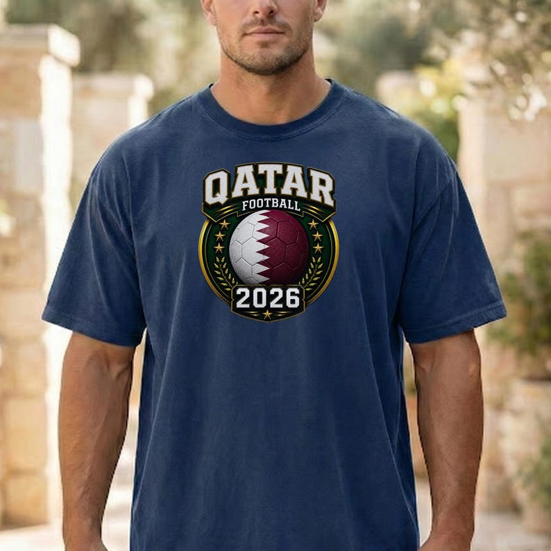 May include: Navy blue t-shirt featuring a Qatar Football emblem. The emblem includes a soccer ball with the Qatar flag design, the text "QATAR FOOTBALL" and the year "2026". The emblem is surrounded by a gold border and green leaves.