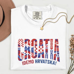 May include: White t-shirt with the word "CROATIA" in red and blue patterned letters. The phrase "IDEMO HRVATSKA!" is printed below. A gold necklace with a star pendant is draped on the shirt.