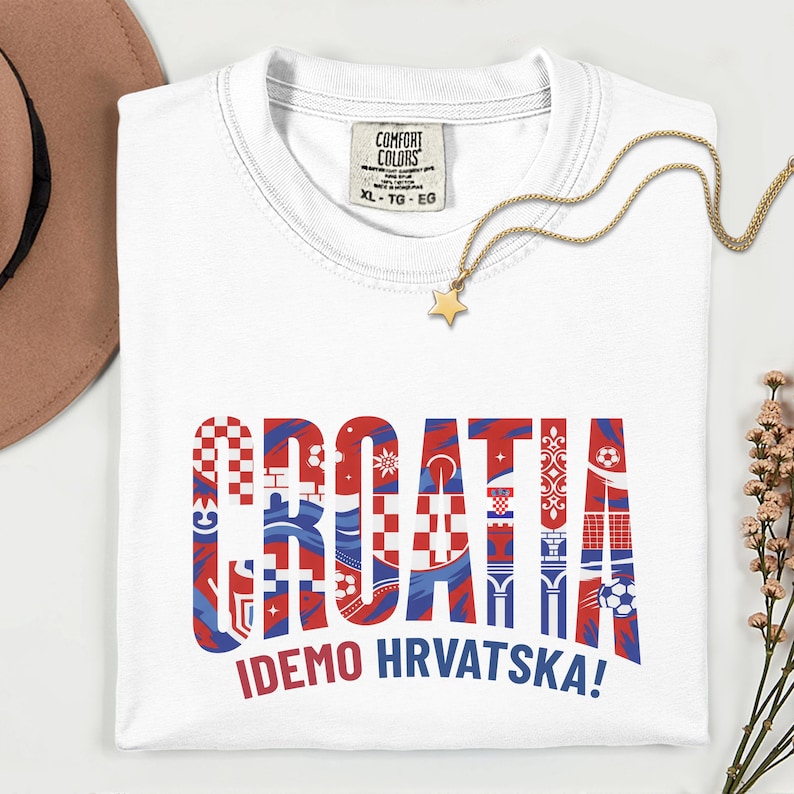 May include: White t-shirt with the word "CROATIA" in red and blue patterned letters. The phrase "IDEMO HRVATSKA!" is printed below. A gold necklace with a star pendant is draped on the shirt.