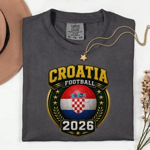 Peut inclure: Un t-shirt gris fonc&eacute; avec un motif de football croate. Le motif comprend le mot "CROATIA" en lettres dor&eacute;es, un ballon de football avec le drapeau croate et l'ann&eacute;e "2026". Un collier dor&eacute; en forme d'&eacute;toile est pos&eacute; sur le t-shirt.