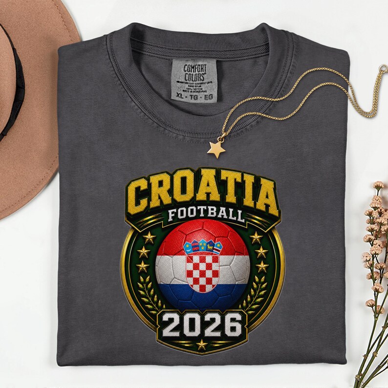 Peut inclure: Un t-shirt gris fonc&eacute; avec un motif de football croate. Le motif comprend le mot "CROATIA" en lettres dor&eacute;es, un ballon de football avec le drapeau croate et l'ann&eacute;e "2026". Un collier dor&eacute; en forme d'&eacute;toile est pos&eacute; sur le t-shirt.