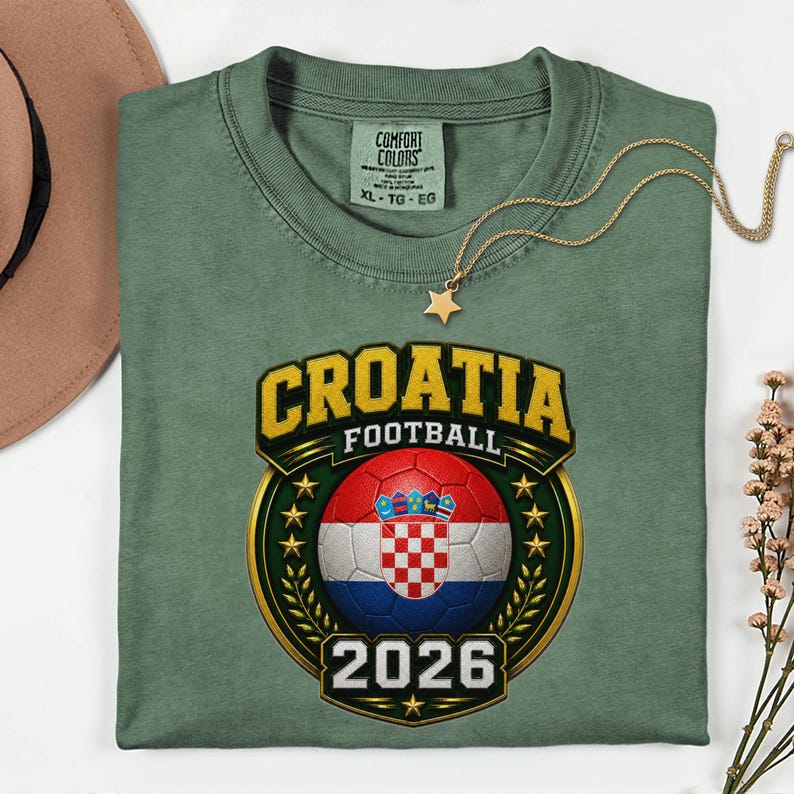 Peut inclure: T-shirt vert sauge avec un motif "Croatia Football". Le motif pr&eacute;sente un ballon de football avec le drapeau croate, le mot "CROATIA" en lettres dor&eacute;es et l'ann&eacute;e "2026". Un collier pendentif &eacute;toile dor&eacute; est drap&eacute; sur le t-shirt.
