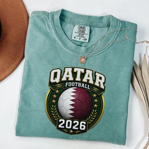 May include: A seafoam green t-shirt featuring a graphic for Qatar Football 2026. The design includes a soccer ball in the colors of the Qatar flag, surrounded by a gold and green emblem. The shirt also has a Comfort Colors tag.