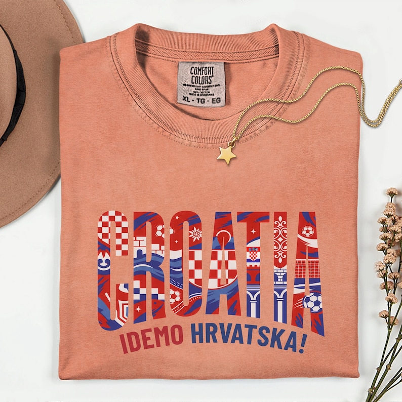 May include: Coral-colored t-shirt with the word "CROATIA" in a decorative, patterned design. The text "IDEMO HRVATSKA!" is below. A gold necklace with a star pendant rests on the shirt.