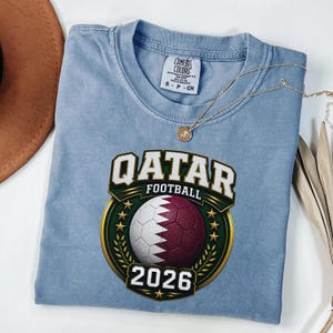 May include: A light blue t-shirt with the text "QATAR FOOTBALL 2026" in white and gold lettering. The shirt features a soccer ball with the Qatar flag design. A gold necklace is draped across the shirt.