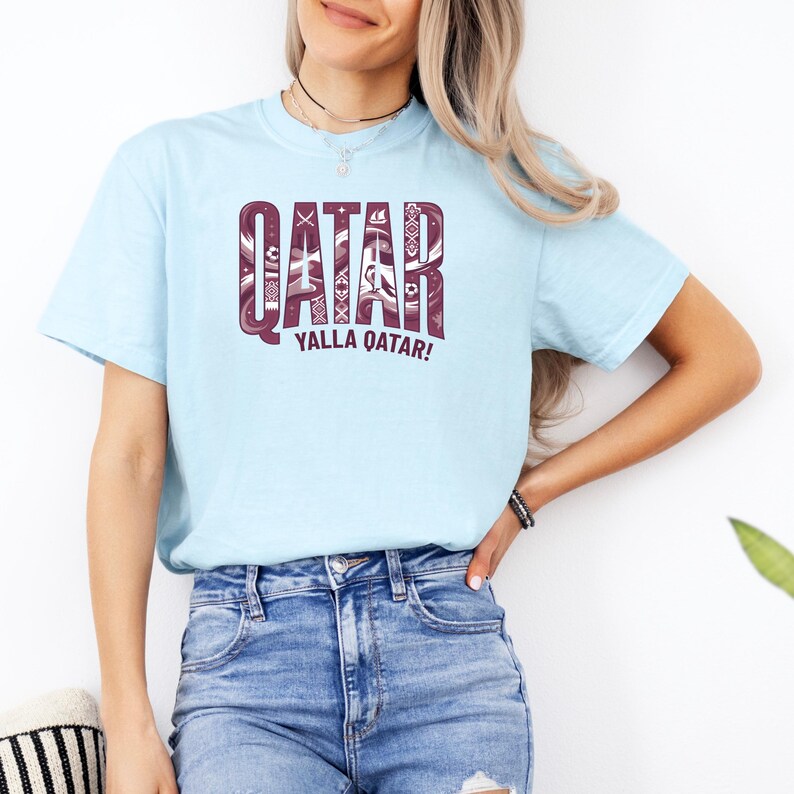 Puede incluir: Una camiseta azul claro con la palabra "QATAR" en un dise&ntilde;o granate estampado. Debajo est&aacute; la frase "YALLA QATAR!" en granate. La camiseta es de cuello redondo.