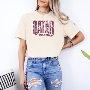 Puede incluir: Camiseta color crema con la palabra "QATAR" en un dise&ntilde;o granate estampado. Debajo, la frase "YALLA QATAR!" en granate. La camiseta se combina con unos vaqueros azules rotos.