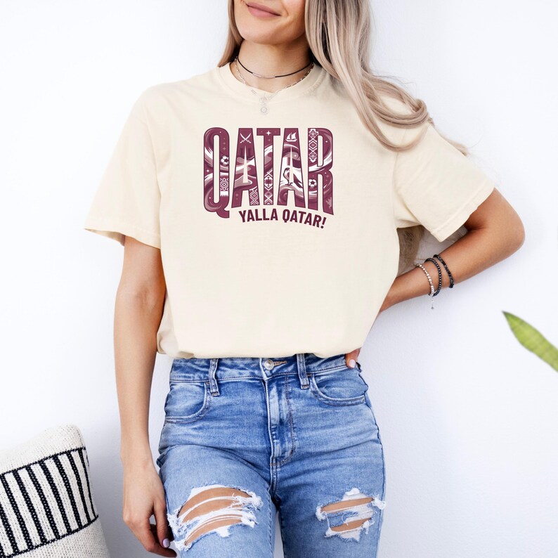 Puede incluir: Camiseta color crema con la palabra "QATAR" en un dise&ntilde;o granate estampado. Debajo, la frase "YALLA QATAR!" en granate. La camiseta se combina con unos vaqueros azules rotos.