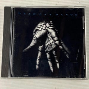 Peut inclure: Un boîtier de CD avec l'album "Dead Can Dance". La pochette présente une image en noir et blanc d'une main à l'aspect squelettique sur fond sombre. Le nom du groupe est imprimé en blanc en haut.