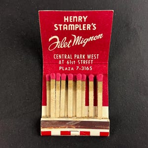 Könnte beinhalten: Ein Vintage-Streichholzbriefchen mit rotem Umschlag. Der Text lautet "HENRY STAMPLER'S Filet Mignon" und die Adresse "CENTRAL PARK WEST AT 61st STREET". Die Streichholzköpfe sind rosa und die Streichhölzer beige.