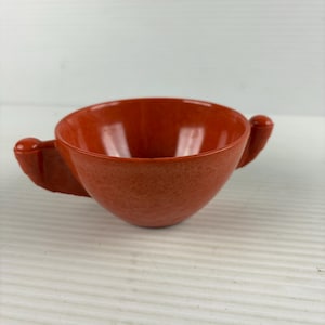 May include: A small, round, orange bowl with a mottled, textured finish. The bowl features two small, rounded handles extending from opposite sides of the rim. The interior is smooth and deep, with a consistent orange color throughout the vessel.