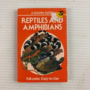 Peut inclure: Un livre Golden Guide intitulé Reptiles and Amphibians avec une couverture orange vif. La couverture présente un serpent à bandes noires et blanches, une petite salamandre orange et une tortue. Un autocollant jaune avec la lettre S est visible.