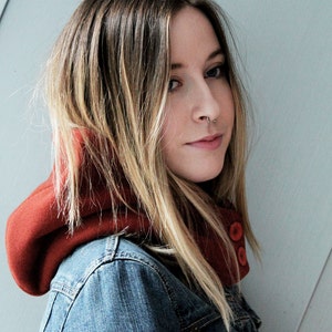 Pode incluir: Uma mulher com cabelo comprido e loiro está usando uma jaqueta de moletom com capuz vermelha com uma jaqueta de jeans por cima. A jaqueta de moletom tem dois botões grandes na frente.