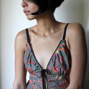 Puede incluir: Un camisola con estampado floral con tirantes finos negros y un frente anudado. La camisola presenta un escote en V y está adornada con encaje negro. La tela tiene un estampado de flores rosas, naranjas y amarillas sobre un fondo gris.
