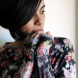 May include: A woman with short black hair is wearing a black long-sleeved shirt with a floral print. The shirt has a colourful trim around the neckline and cuffs.