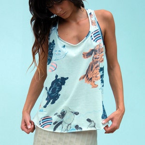 May include: A woman wearing a light blue tank top with a print of dogs and cats playing with balls. The tank top has a scoop neck and a racerback style. She is also wearing a light beige skirt.