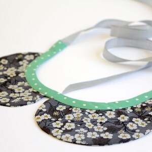 Puede incluir: Un collar de tela floral gris y blanco con un ribete verde de lunares. El collar está unido a una cinta gris.