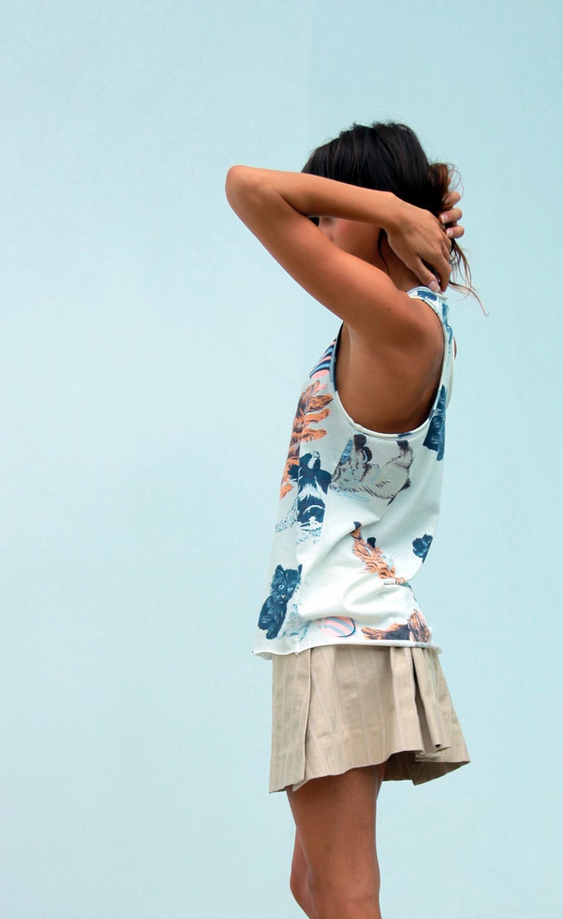 May include: A woman wearing a white tank top with a colorful cat print and a beige pleated skirt.