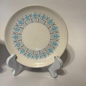 Op de afbeelding: Crèmekleurige keramische plaat met een blauw-grijs bloemmotief langs de rand. De plaat wordt gepresenteerd op een witte houten standaard. Een vintage stuk, geschikt voor het serveren van eten of als decoratie.