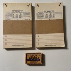 May include: Two identical vintage 1953 daily tear-off calendars with cream paper, black text, and a brown paper band, showing January 1st, Thursday. Below them is a small rectangular box labeled 100 Steel City No. 1 Gem Paper Clips.