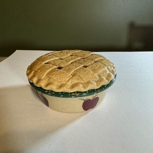 Op de afbeelding: Een keramische taartvorm met een deksel van een roosterkorst. De schaal heeft een beige basis met groene rand en paarse appelontwerpen. Het taartkorstdeksel is lichtbruin met een textuur, geweven patroon. De schaal is rond.