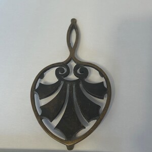 May include: An antique-style trivet with a dark bronze finish. The trivet has a stylized leaf design and a handle. The metalwork is detailed and decorative, suitable for kitchen or dining decor. Measures approximately 18 cm in length.