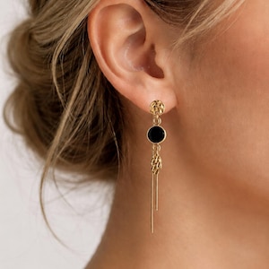 Peut inclure: Une boucle d'oreille pendante dorée composée d'un clou circulaire texturé, d'une breloque circulaire noire lisse, d'un maillon ovale texturé et de deux fines chaînes pendantes. Le bijou présente plusieurs segments reliés créant un design vertical.