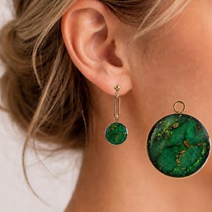Op de afbeelding: Goudkleurige oorbellen met een cirkelvormig, groen en goud gemarmerd ontwerp. Eén oorbel is een knop met een rechthoekige druppel, de andere is een grote hanger. De oorbellen hebben een natuurlijke steenlook.