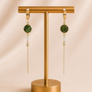 Op de afbeelding: Goudkleurige oorbellen met groene, cirkelvormige stenen en bungelende kettingen. De oorbellen worden gepresenteerd op een gouden sieradenstandaard tegen een zachte, neutrale achtergrond. De oorbellen hebben een delicaat ontwerp met kleine, heldere accenten.