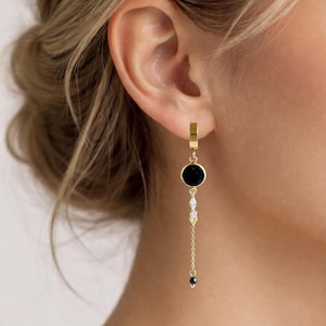 Op de afbeelding: Een gouden hangende oorbel met een kleine ring aan de bovenkant, een ronde zwarte steen, twee kleine heldere marquise-geslepen stenen, een fijne gouden ketting en een minuscuul donkerblauw kraaltje onderaan, verticaal gerangschikt.