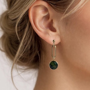 Op de afbeelding: Goudkleurige oorhangers. Elke oorbel heeft een kleine stud, een verticale ovale schakel en een ronde hanger met een donkergroen en blauw gemarmerd ontwerp. De oorbellen zijn te zien aan een oor.