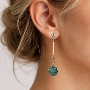 Op de afbeelding: Een goudkleurige hangende oorbel bestaande uit een kleine bolvormige knop verbonden met een verticale staaf, een cirkelvormige schakel en een ronde hanger met een textuurrijk, gevlekt blauwgroen oppervlak dat verticaal omlaag hangt.