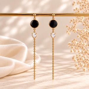 Op de afbeelding: Een paar goudkleurige oorbellen met een ronde zwarte steen aan de bovenkant, gevolgd door een heldere peervormige kristal en eindigend met een lange, fijne goudkleurige ketting. De oorbellen hangen aan een dunne horizontale goudkleurige staaf.