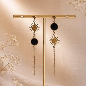 Op de afbeelding: Goudkleurige hemelse oorbellen met een zonnesterontwerp en een zwart cirkelvormig accent. De oorbellen hangen aan een ketting en worden tentoongesteld op een gouden standaard. De achtergrond is een zachte, neutrale kleur.