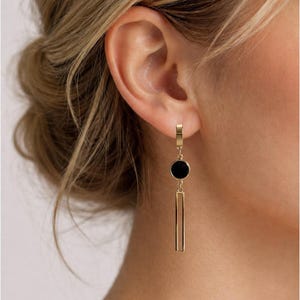 Op de afbeelding: Goudkleurige oorhangers met een zwarte cirkelvormige accent en een rechthoekige staaf. De oorbellen worden getoond tegen de achtergrond van een oor en haar, wat het ontwerp benadrukt. Een stijlvol accessoire.