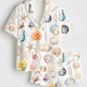 Puede incluir: Conjunto de pijama blanco con estampado de conchas marinas. La camisa de manga corta con botones y los pantalones cortos a juego están cubiertos de ilustraciones coloridas de varias conchas, estrellas de mar y coral.