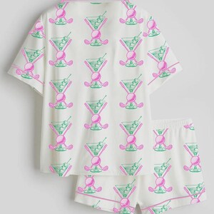 May include: White pajama set with a repeating pattern of pink golf clubs, golf balls, and green martini glasses. The set includes a short-sleeved button-up shirt and matching shorts, both trimmed with pink piping.