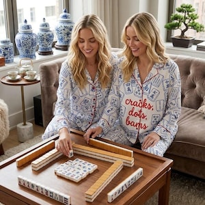 May include: Two women wearing matching white pajamas with blue and red accents, playing Mahjong on a wooden table. The pajamas have a Mahjong tile pattern and the words "craicks, dots, bams" in red.