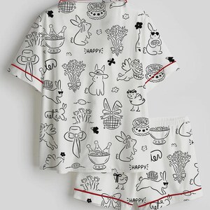 以下が含まれることがあります： 白地に黒と赤のトリムが付いたパジャマセット。半袖トップスとショートパンツのセットで、ウサギ、花、ケーキ、そして「Happy」の文字が黒い線画で描かれています。