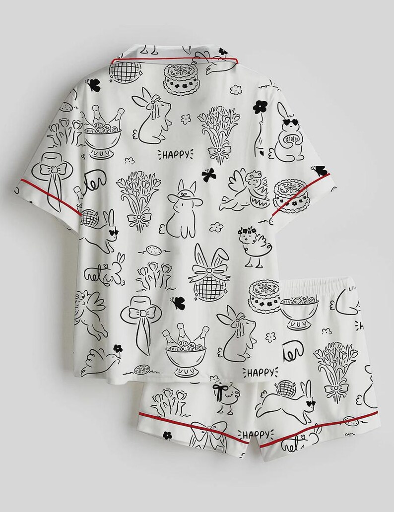 以下が含まれることがあります： 白地に黒と赤のトリムが付いたパジャマセット。半袖トップスとショートパンツのセットで、ウサギ、花、ケーキ、そして「Happy」の文字が黒い線画で描かれています。
