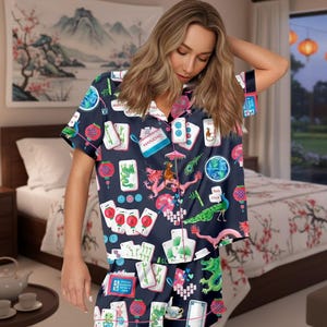 K&ouml;nnte beinhalten: Marineblaues Pyjama-Set mit Mahjong-Kachel-Print mit Drachen, Laternen und Kartensymbolen. Das kurz&auml;rmelige Oberteil und die Shorts haben einen rosa Besatz. Der Hintergrund umfasst ein Schlafzimmer mit einer floralen Tagesdecke und einem Landschaftsgem&auml;lde.
