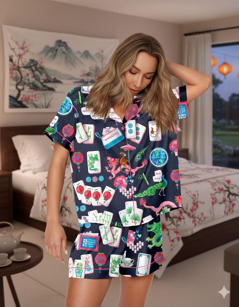 K&ouml;nnte beinhalten: Marineblaues Pyjama-Set mit Mahjong-Kachel-Print mit Drachen, Laternen und Kartensymbolen. Das kurz&auml;rmelige Oberteil und die Shorts haben einen rosa Besatz. Der Hintergrund umfasst ein Schlafzimmer mit einer floralen Tagesdecke und einem Landschaftsgem&auml;lde.