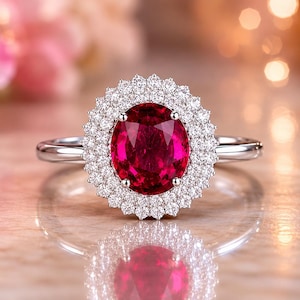 Peut inclure: Une bague en argent ornée d'une grosse pierre rouge vif de forme ovale, entourée d'un halo de petites pierres claires. La bague est posée sur un fond flou aux tons roses et dorés.
